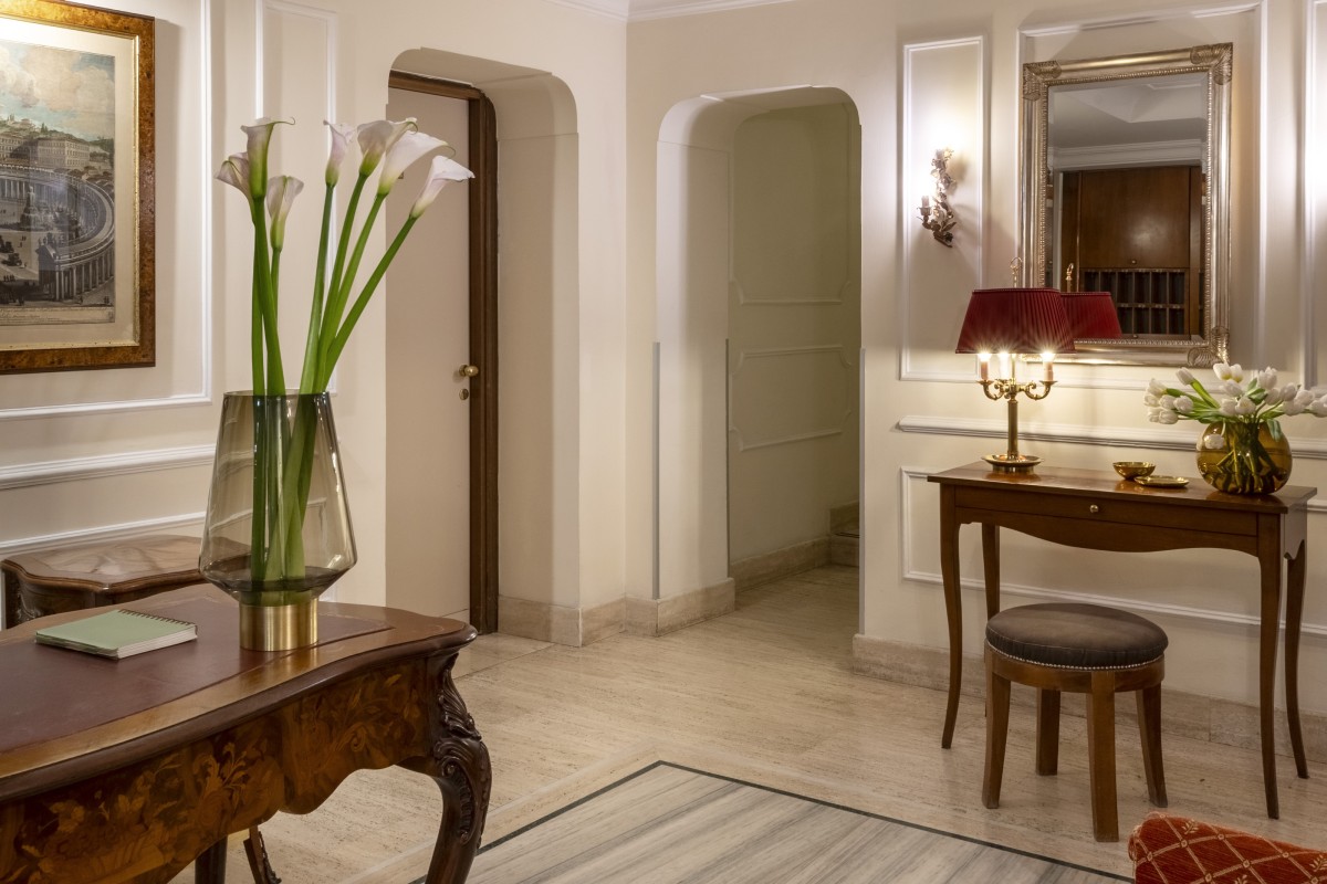 Elegant lobby of Hotel Cortina in Rome, featuring an antique wooden desk, a vase of flowers, an ornamental mirror, and polished marble floors - Hotel Cortina, BZAR hotels in Rome