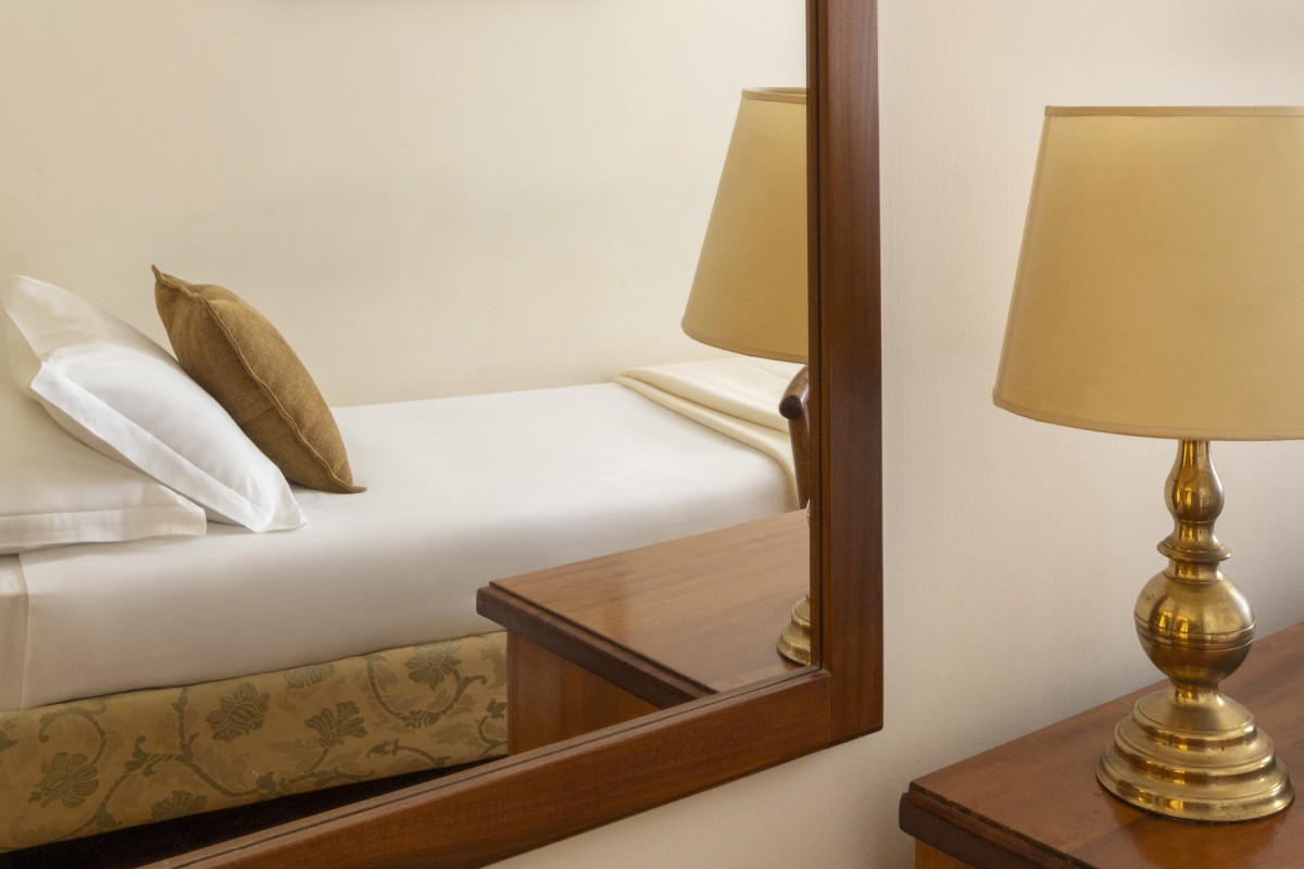 Details of a single room at Hotel Cortina in Rome, featuring classic decor with a wooden desk, vintage chair, suitcase by the window, and a single bed with crisp white linens - Hotel Cortina, BZAR hotels a Roma