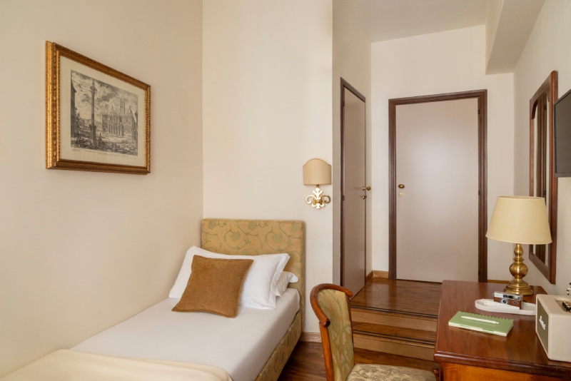 Cozy single room interior with parquet flooring, single bed with patterned headboard, wooden desk with a classic lamp, and entrance door - Hotel Cortina, BZAR hotels a Roma