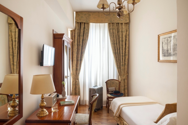 Bright single room at Hotel Cortina in Rome, featuring classic decor with a wooden desk, vintage chair, suitcase by the window, and a single bed with crisp white linens - Hotel Cortina, BZAR hotels a Roma