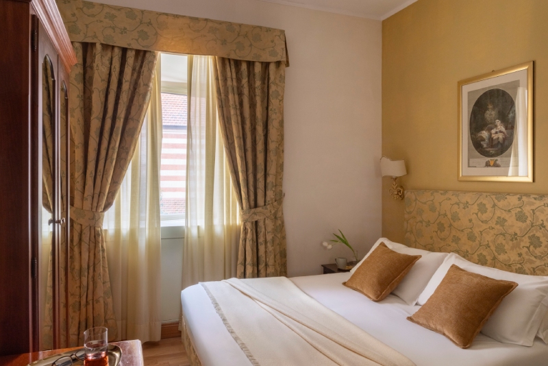 Cozy detail of a well-organized Small room at Hotel Cortina in Rome, showing a single bed, a period desk, an upholstered chair, and a framed mirror reflecting window light - Hotel Cortina, BZAR hotels in Rome