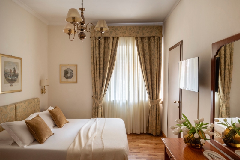 Welcoming standard room at Hotel Cortina in Rome, featuring a comfortable double bed with brown pillows, parquet flooring, and a large window with elegant floral curtains - Hotel Cortina, BZAR hotels in Rome