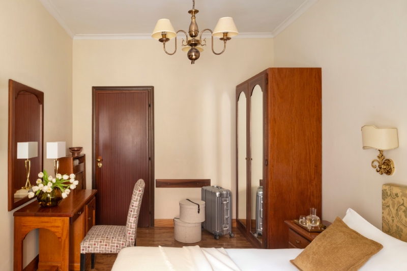 Spacious interior of a standard room at Hotel Cortina in Rome, featuring an antique wooden desk, an upholstered chair, a large built-in wardrobe, and a silver trolley suitcase - Hotel Cortina, BZAR hotels in Rome